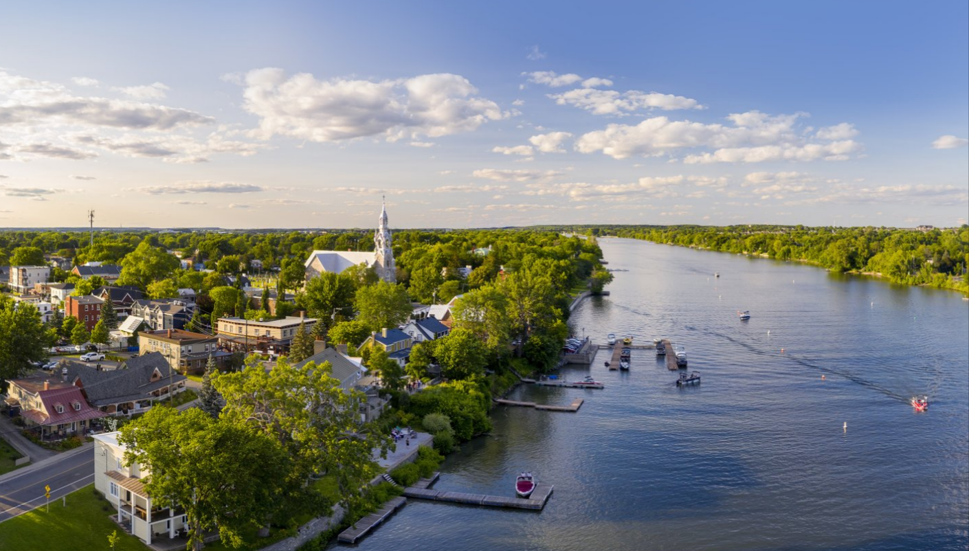 Vue aérienne d’un village en Montérégie qui borde la rivière Richelieu. On y voit une église au clocher blanc entourée d’arbres verdoyants, des maisons colorées, un quai avec des bateaux amarrés, et un ciel bleu parsemé de nuages.